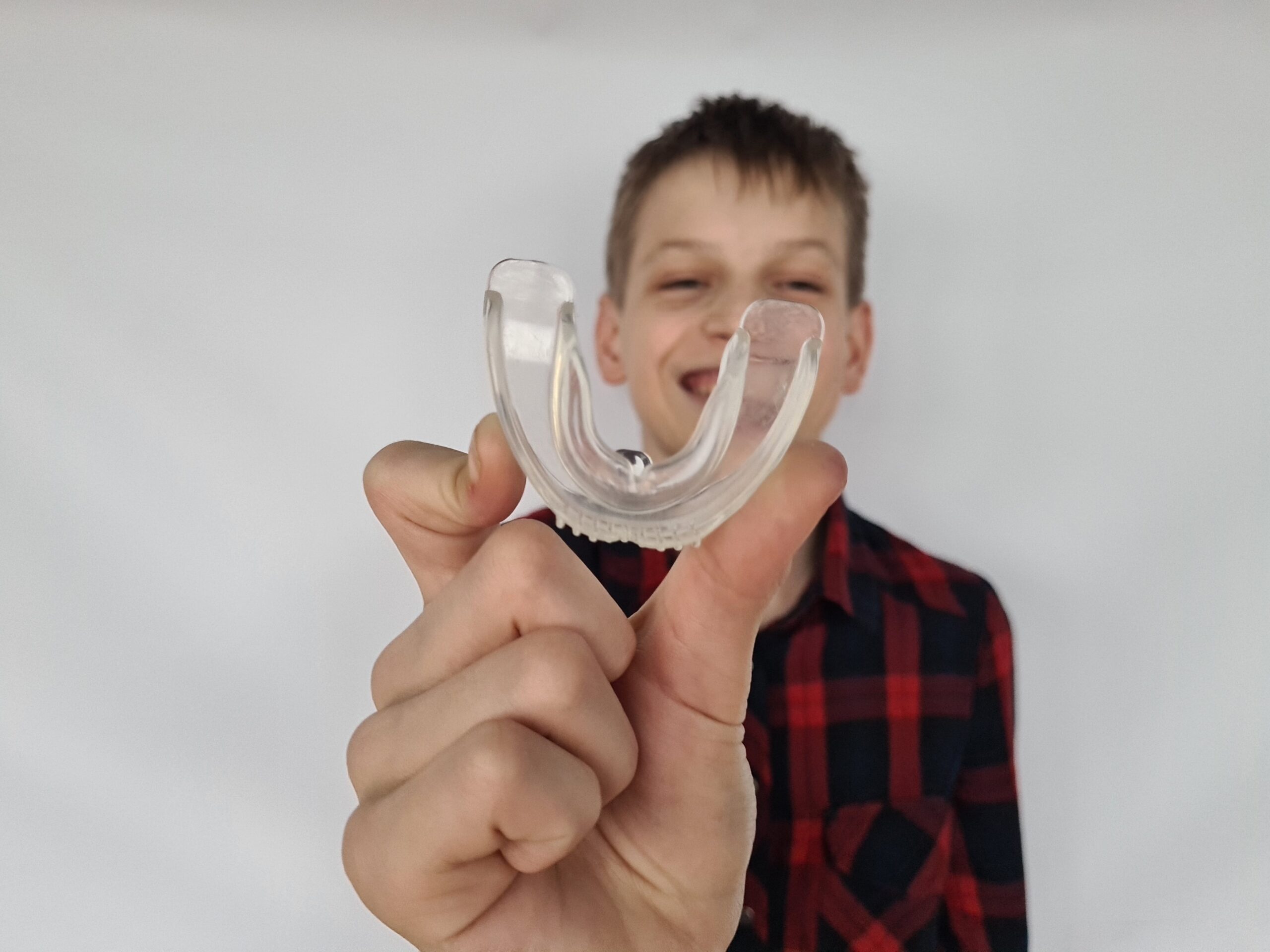 A boy smiles while holding a clear mouthguard in front of a plain background, illustrating dental care and sports safety