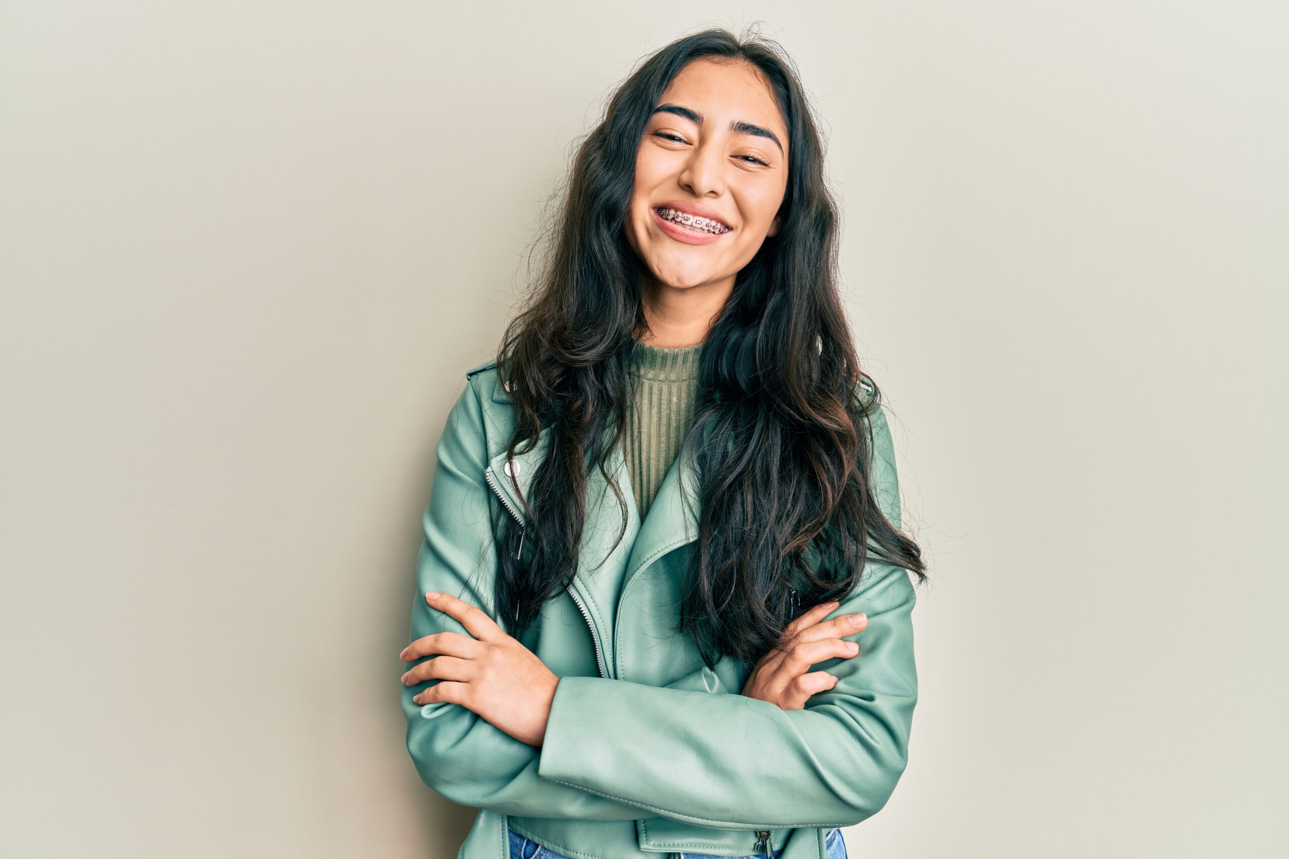 Hispanic teenager girl with dental braces wearing green leather jacket happy face smiling with crossed arms looking at the camera. positive person.